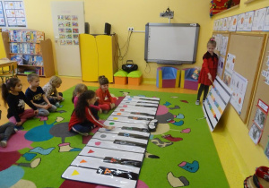 Dziewczynka wskazuje kolejność granych dźwięków na tablicy muzycznej, trzy dziewczynki odtwarzają dźwięki na instrumencie.